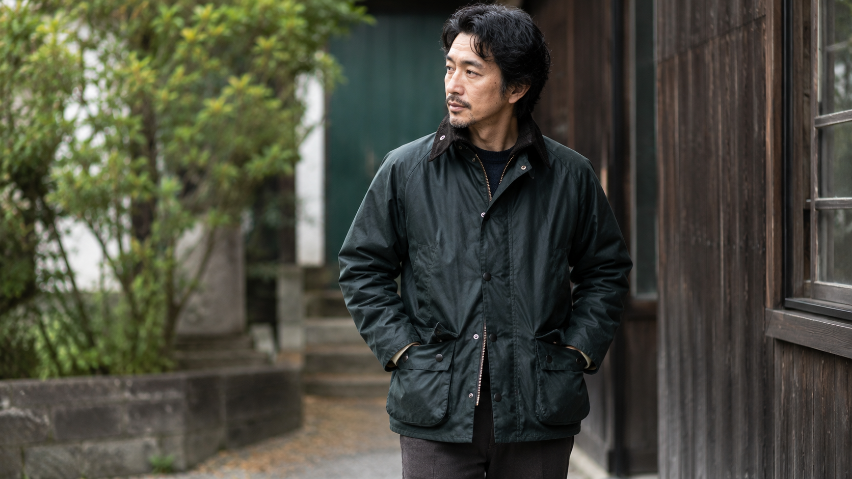 バブアーのオイルジャケットを着た40代男性のコーディネート。クラシックな木造背景で落ち着いた大人のヴィンテージスタイルを演出している。