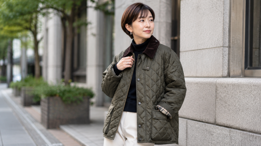 バブアーのキルティングジャケットを着た40代女性のコーディネート。都会的な街並みを背景に、落ち着いた大人のカジュアルスタイルを演出している。
