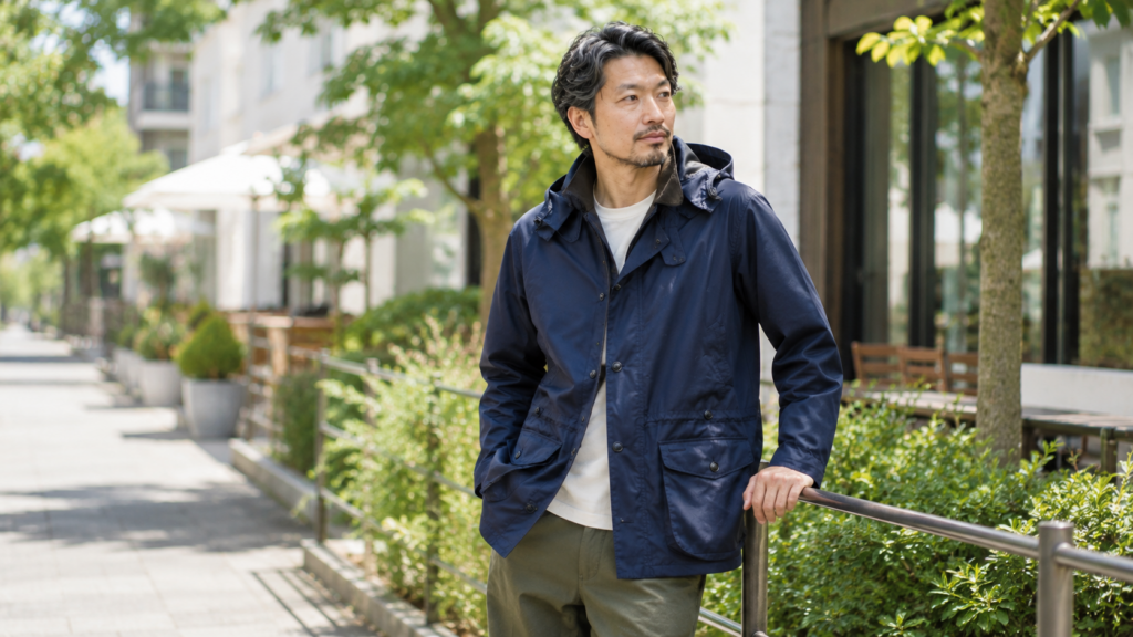 春夏らしい街並みを背景に、ネイビーのバブアージャケットを着た40代男性のコーディネート。爽やかな緑と自然光が映える大人のカジュアルスタイル。